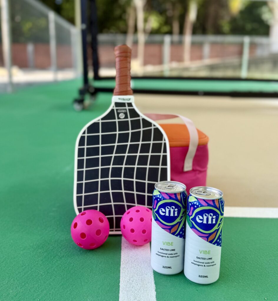 A pickleball racket, two hot-pink pickleballs, two cans of effi VIBE functional soda and a pink and orange cooler bag on a pickleball court
