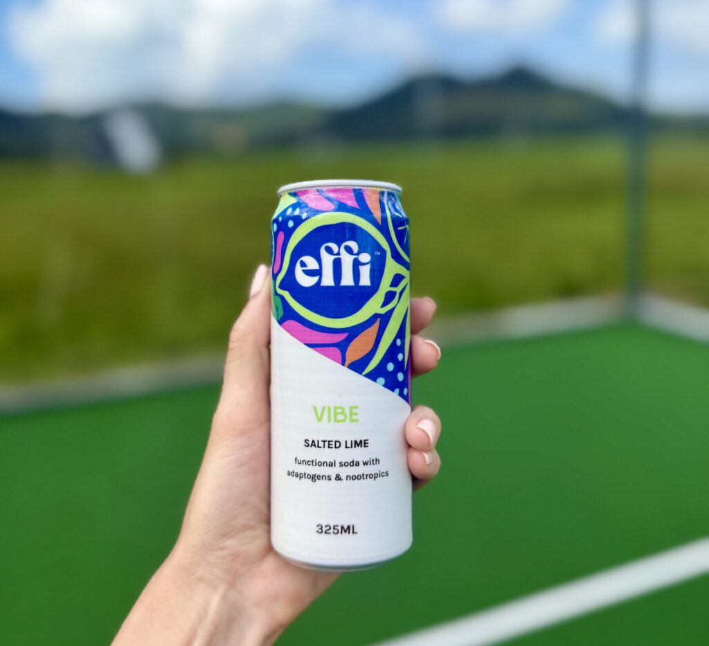 A hand with light pink painted nails holding a can of effi VIBE functional soda on a padel court, with a rolling green landscape behind